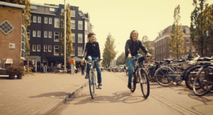 Deux adolescents souriants faisant du vélo côte à côte sur une large rue pavée d'Amsterdam sous une lumière dorée et chaude de fin d'après-midi, sans obstruction centrale.