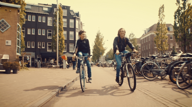 Deux adolescents souriants faisant du vélo côte à côte sur une large rue pavée d'Amsterdam sous une lumière dorée et chaude de fin d'après-midi, sans obstruction centrale.