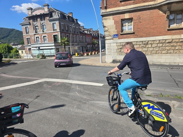 Autre usager en t-shirt bleu marine et pantalon bleu vif roulant sur un vélo électrique Vélo Fluo dans les rues de Revin, illustrant la simplicité de l'offre.