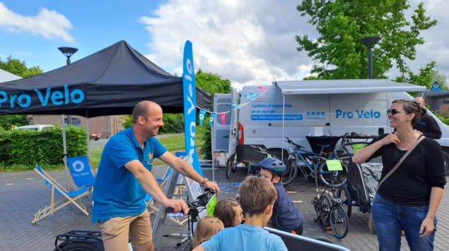 Membre de l'équipe Pro Velo sur un vélo cargo, entouré d'enfants, devant le stand et le camion Pro Velo
