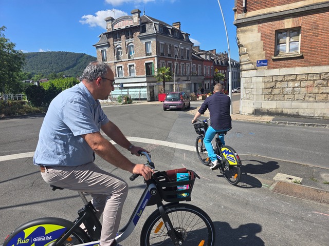 Homme en chemise bleue et pantalon beige roulant sur un vélo à assistance électrique Vélo Fluo à Revin, près de la gare.