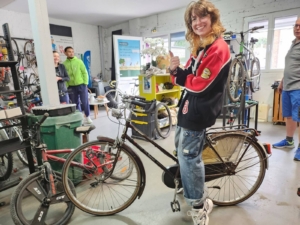 Jeune femme souriante levant le pouce à côté d’un vélo de ville, dans un atelier vélo rempli de vélos.