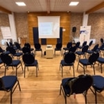 Vue d’ensemble d’une salle sous charpente en bois, avec le public assis face à l’écran de présentation.