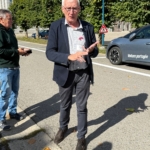 Homme en veste sombre donnant des explications en extérieur, près de voitures garées.