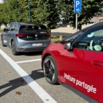 Voiture électrique grise garée à côté d’une voiture rouge, sur un parking en extérieur.